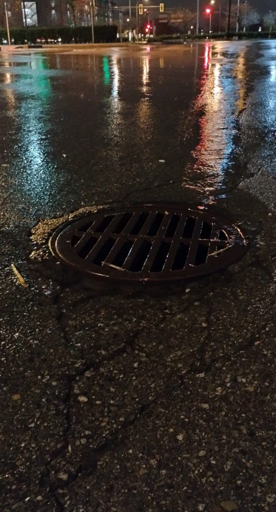 Night picture of a storm drain surrounded by puddles reflecting traffic lights.